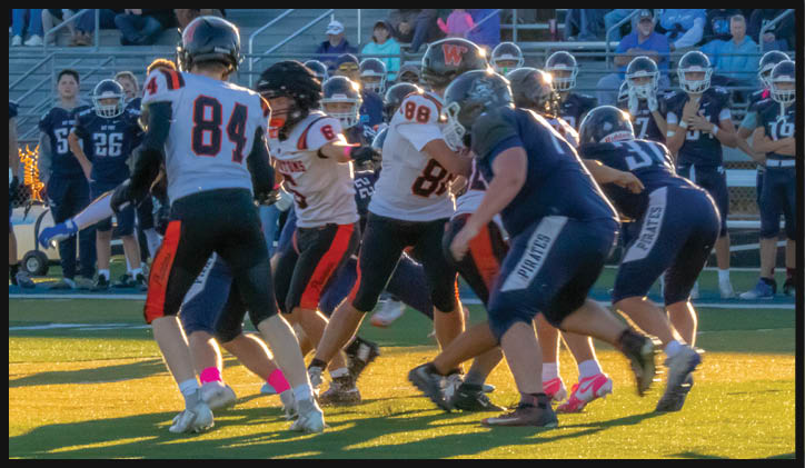 People wearing football uniforms are actively engaged in a game on a field with spectators seated in bleachers in the background. Description generated by AI