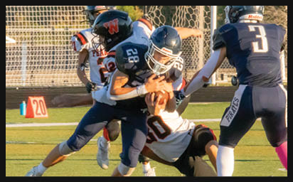 A person wearing a dark blue football uniform with the number 28 is holding a football tightly while being tackled by two people in white and orange uniforms on a grassy field. Description generated by AI
