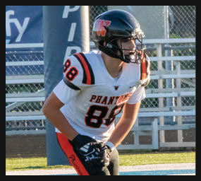 A person wearing a football helmet and a white jersey with the number 88 is standing on a field near a goalpost and bleachers. Description generated by AI