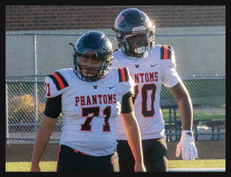 Two people wearing football helmets and white jerseys with “PHANTOMS" written on them stand on a field with a chain-link fence and brick wall in the background. Description generated by AI