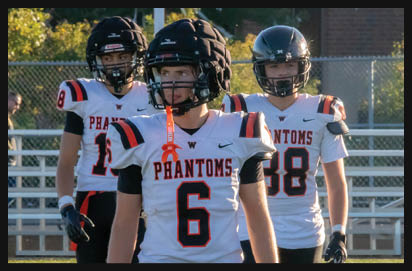 Three people wearing football helmets and white jerseys with “Phantoms" written on them are standing on a field with bleachers and trees in the background. Description generated by AI