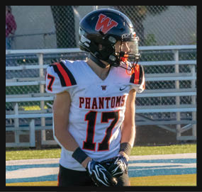 A person wearing a football uniform with the number 17 and a helmet stands on a field near bleachers. Description generated by AI
