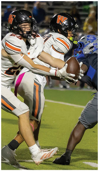 Two people in football uniforms are engaged in a play on a field with one holding the ball while a third player in a different uniform attempts to tackle. Description generated by AI