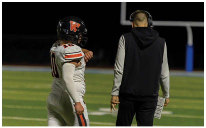 A person in a football uniform with the number 10 is standing next to a person wearing a headset and holding a play sheet on a football field. Description generated by AI