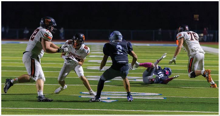 Four people in football uniforms are on a field with one person in dark clothing falling backward while the others in white uniforms approach. Description generated by AI