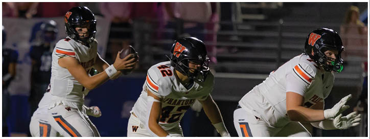 Three people in football uniforms are on a field with one holding a football preparing to pass while the others are in ready positions. Description generated by AI