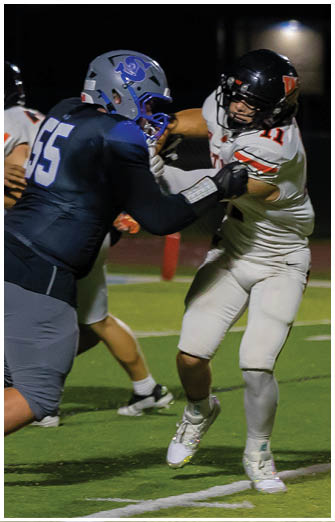 Two people in football uniforms are engaged in a play on a field under stadium lights, with one person wearing a dark blue jersey and helmet and the other in a white jersey and black helmet. Description generated by AI