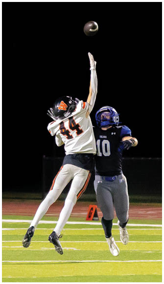 Two people in football uniforms are jumping on a field at night reaching for a football in midair. Description generated by AI