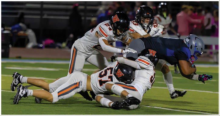 Three people in football uniforms are engaged in a play on a field, with two players in white uniforms tackling a player in a dark uniform who is holding the ball. Description generated by AI