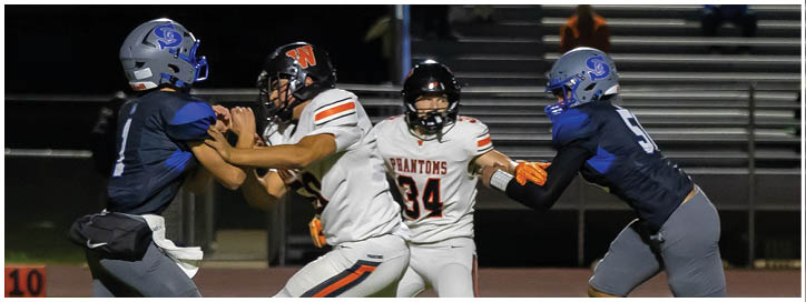 Three people in football uniforms are engaged in a play on a field with one person in white running between two people in blue who are attempting to block or tackle. Description generated by AI