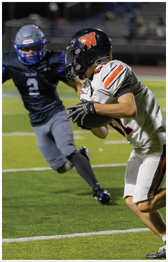 Two people in football uniforms are engaged in a play on a field, with one person holding the ball and the other reaching out to tackle. Description generated by AI