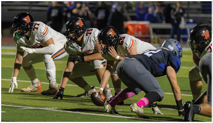 People in football uniforms are lined up on a field preparing for a play during a game. Description generated by AI