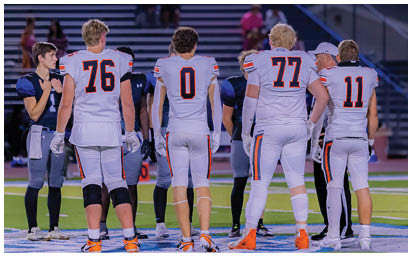 Four people wearing white sports jerseys with numbers 76, 0, 77, and 11 stand on a field facing away from the camera during sunset with a stadium and spectators in the background. Description generated by AI