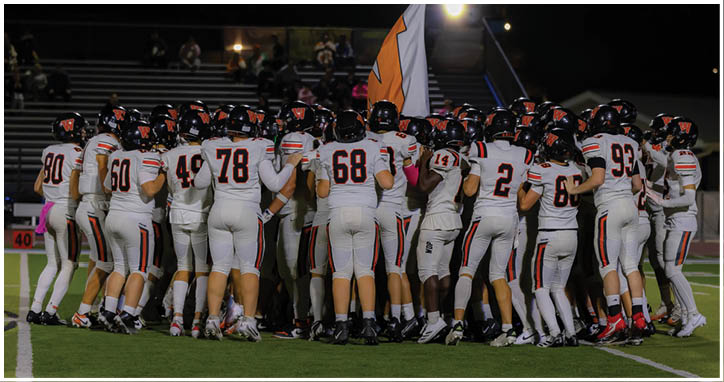 A group of football players in white uniforms with black helmets are gathered closely together on a field at night with a flag visible in the background. Description generated by AI