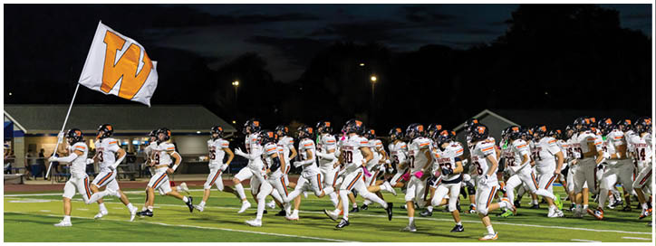 A large group of people wearing white sports uniforms with helmets is running together on a green football field at night. Description generated by AI