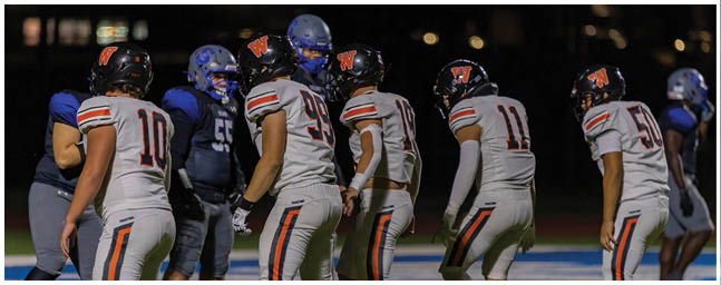 Several people wearing white football uniforms with orange and black stripes are standing on a green field, preparing for a play. Description generated by AI