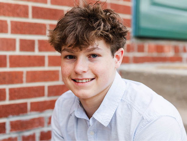 A person with tousled hair and a light-colored shirt is smiling in front of a brick wall. Description generated by AI