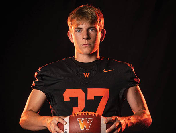 A person with short hair wearing a black sports jersey with the number 27 is holding a football with a “W" logo against a dark background. Description generated by AI