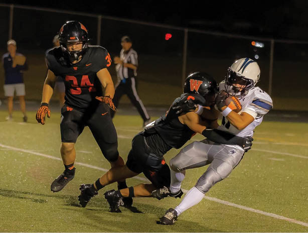 Two people in black football uniforms tackle a person in a white and gray uniform holding a football on a lit field during a night game. Description generated by AI