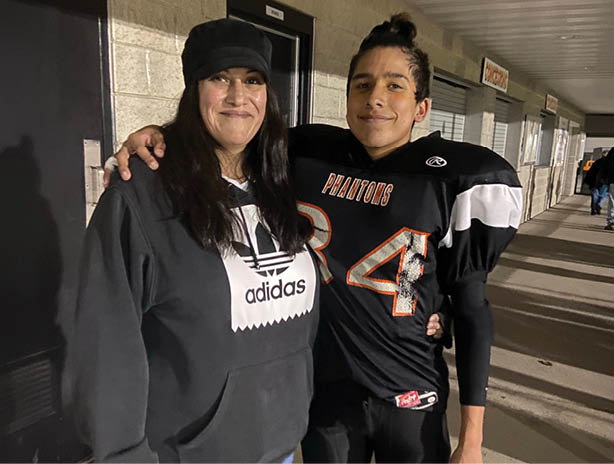 A person wearing a black hat and jacket stands smiling next to a person in a football uniform with shoulder pads inside a building hallway. Description generated by AI