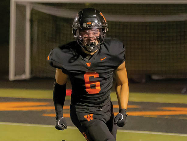 A person wearing a black football uniform with the number six in orange is running on a field with a goalpost in the background. Description generated by AI