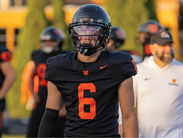 A person wearing a black football uniform with the number six and a helmet stands on a field with other people and trees in the background. Description generated by AI