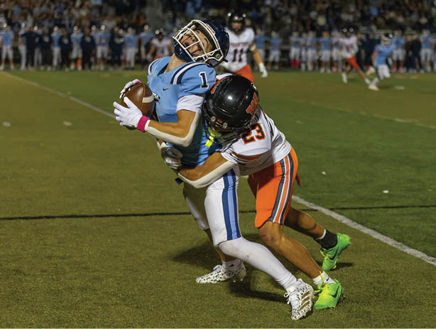 A person in a light blue sports uniform is being tackled by another person in an orange and white uniform during a football game on a grassy field. Description generated by AI