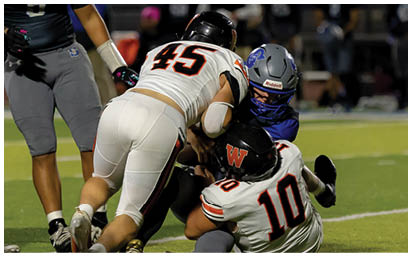 Three people in football uniforms are engaged in a tackle on a grassy field during a game. Description generated by AI