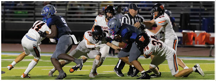 Several football players in helmets and uniforms are engaged in a tackle on a field with a referee observing nearby and spectators in the stands. Description generated by AI