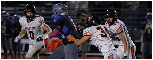 People wearing football uniforms are actively playing on a field with one person in dark gear running while others in white uniforms attempt to tackle. Description generated by AI