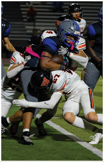 A group of people in football uniforms are engaged in a tackle on a field during a game, with one person holding the ball tightly while others attempt to stop them. Description generated by AI