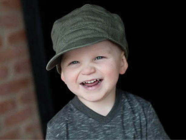 A smiling child wearing a green cap and a dark gray t-shirt stands in front of a dark background with a brick wall partially visible. Description generated by AI