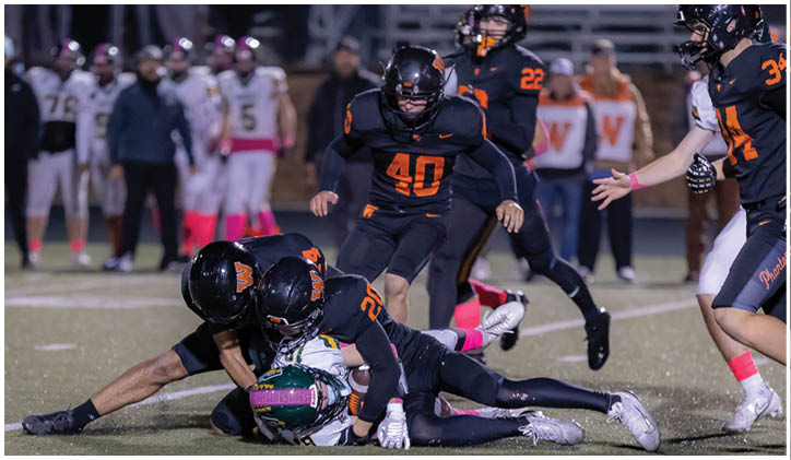 Several people in dark sports uniforms are tackling a person in a white and green uniform on a football field during a game. Description generated by AI