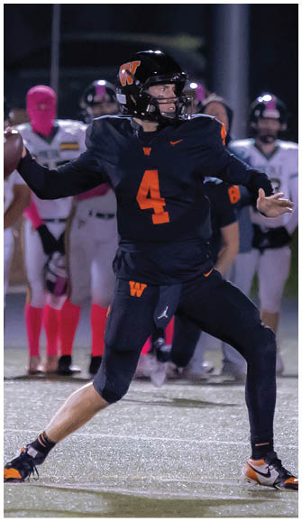 A person in a dark football uniform with the number 4 is preparing to throw a football on a field while several people in white uniforms stand in the background. Description generated by AI