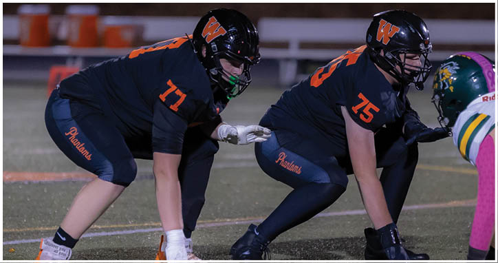 Two people in dark sports uniforms with helmets are crouched in a ready position on a football field facing an opponent in a green and yellow uniform. Description generated by AI