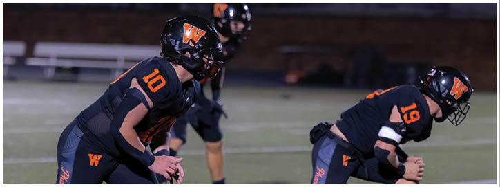 Three people in black football uniforms with orange accents are positioned on a field, appearing ready for a play. Description generated by AI
