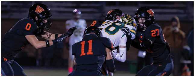 Three people in football uniforms are engaged in a play on a field, with two players in dark uniforms surrounding a player in a white and green uniform holding the ball. Description generated by AI