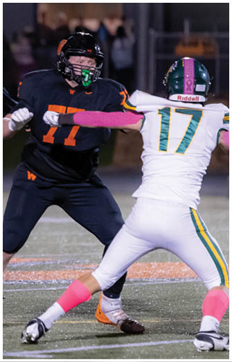 Two people in black football uniforms with orange accents are actively engaged in a play on a football field at night. Description generated by AI