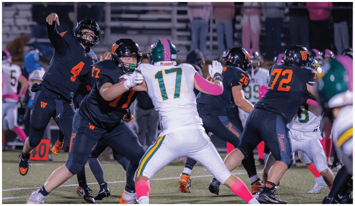 Adults in football uniforms are actively playing on a field with one player throwing a ball while others block and defend. Description generated by AI