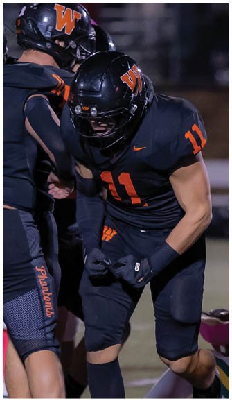 Three people wearing black football uniforms with orange numbers and helmets are engaged in a play on a football field at night. Description generated by AI