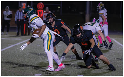 A group of people in football uniforms are actively playing on a field at night with one person holding the ball and others attempting to tackle or block. Description generated by AI