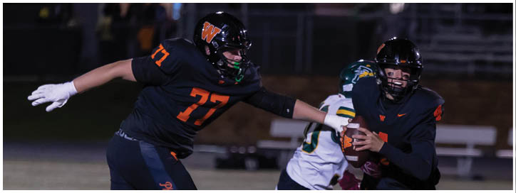 Two people in football uniforms are engaged in a play on a field at night, with one person holding the ball and the other reaching out to block or tackle. Description generated by AI