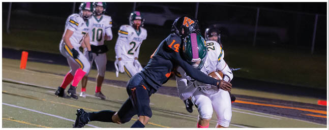 Two people in football uniforms are engaged in a tackle on a field while other players stand in the background. Description generated by AI