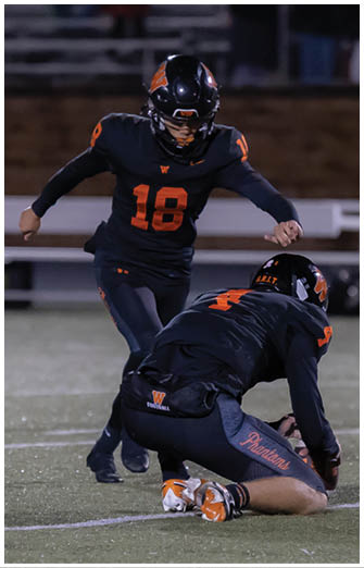 Two people in black and orange football uniforms are on a field with one person kneeling and holding a football while the other prepares to kick it. Description generated by AI