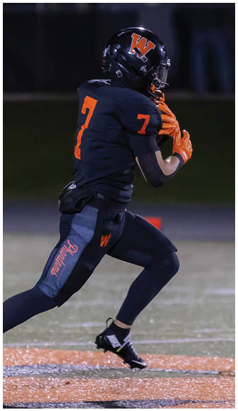 A person wearing a black football uniform with orange numbers and gloves is running on a field. Description generated by AI