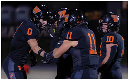 Two people wearing black football uniforms with orange numbers and helmets are bumping fists on a field at night. Description generated by AI
