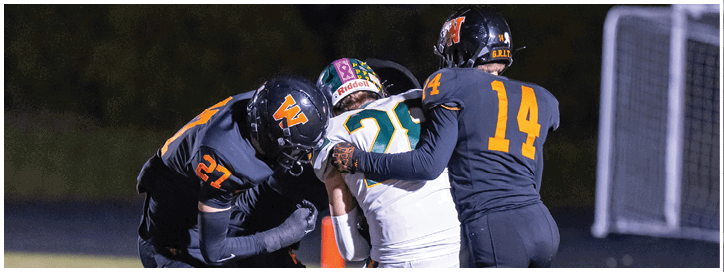 Three people in football uniforms are engaged in a tackle on a field near a goalpost. Description generated by AI