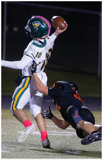 A person in a white football uniform is throwing a football while another person in a black uniform is diving to tackle them on a lit football field at night. Description generated by AI