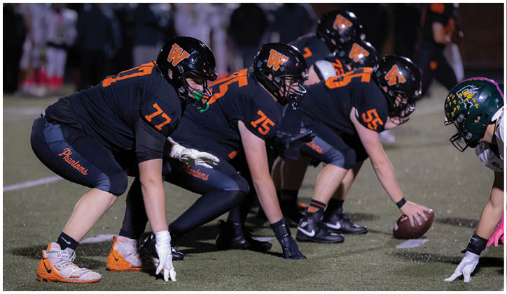 Several people in football uniforms are lined up on a field in a ready stance facing an opponent preparing to snap the ball. Description generated by AI