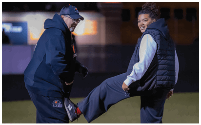 Two people dressed in athletic clothing are stretching on a field at night, with one person holding the other's foot against their thigh. Description generated by AI
